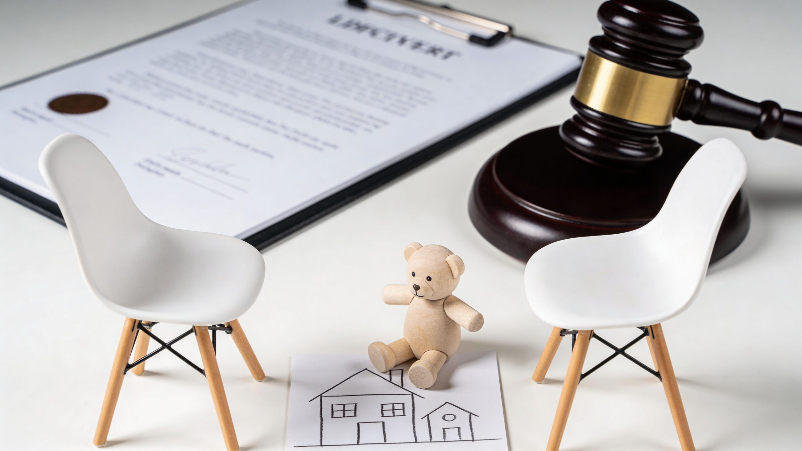 A gavel, a legal document, and a toy bear sitting between two small chairs with a house drawing.