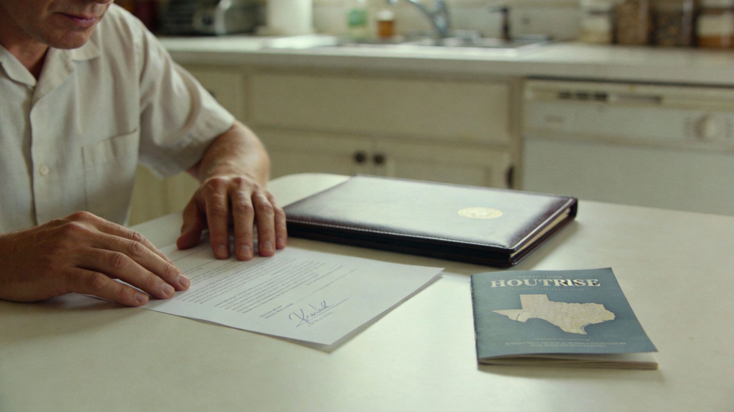 A man sits at a kitchen table reviewing a legal document next to a folder and booklet.