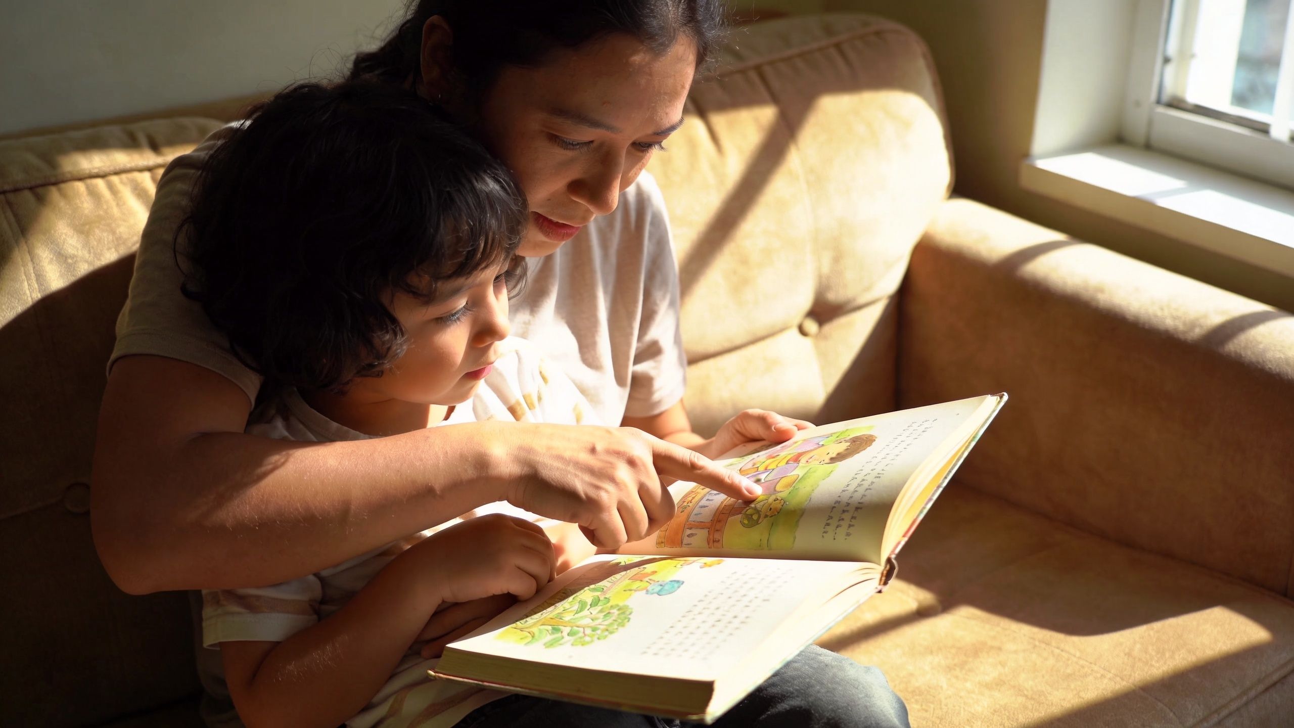 A mother sitting on a couch with her child, reading an open storybook together in natural sunlight.